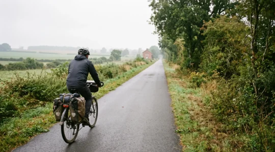 Un cycliste à vélo avec sacoches roule sur la voie verte normande aménagée sur une ancienne ligne ferroviaire, entre arbres et plateaux agricoles.