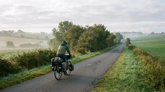 Un cycliste pédale en solitude sur une voie verte arborée entre les haies du bocage normand, en lumière matinale douce.