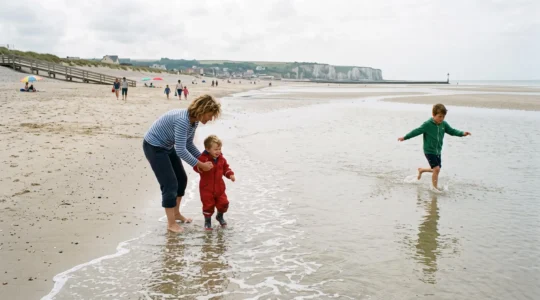 Des enfants pataugent dans une eau peu profonde sur une plage de sable en pente douce en Normandie, accompagnés de leurs parents.