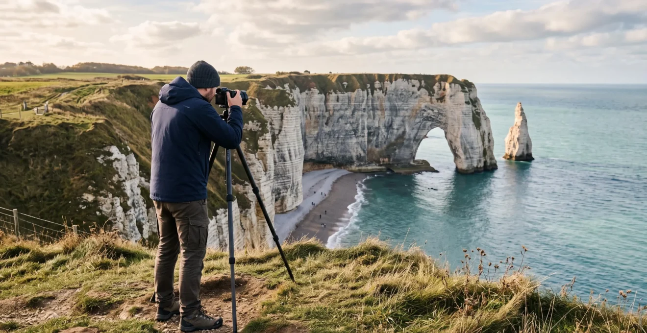 Un photographe au trépied compose une image depuis le plateau herbeux de la falaise d'Aval à Étretat, face à l'arche de craie blanche au-dessus de la mer.
