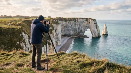 Un photographe au trépied compose une image depuis le plateau herbeux de la falaise d'Aval à Étretat, face à l'arche de craie blanche au-dessus de la mer.
