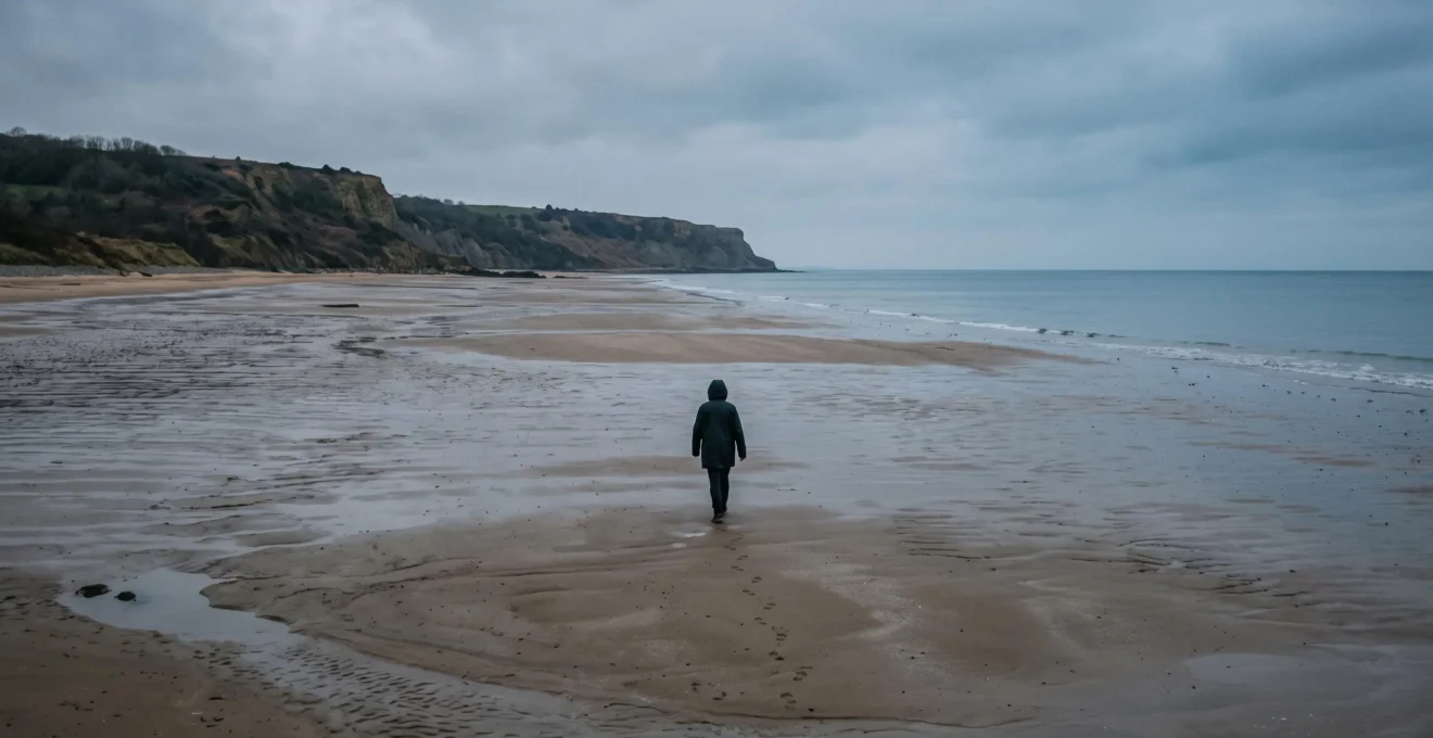 Un visiteur marche seul à marée basse sur la plage d'Omaha, en Normandie, sous un ciel couvert, face aux falaises au loin.