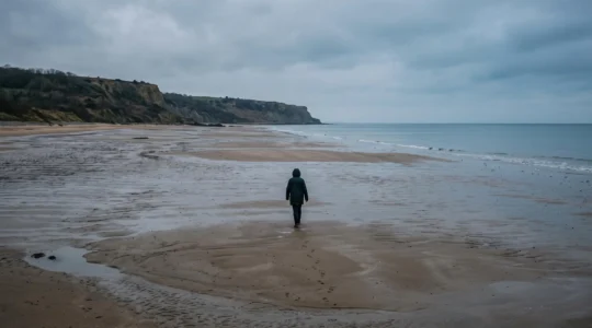 Un visiteur marche seul à marée basse sur la plage d'Omaha, en Normandie, sous un ciel couvert, face aux falaises au loin.